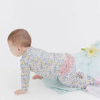 A baby crawling on a white studio floor wearing a long-sleeve footed onesie with a dense pastel floral print and a pink ruffled back detail.