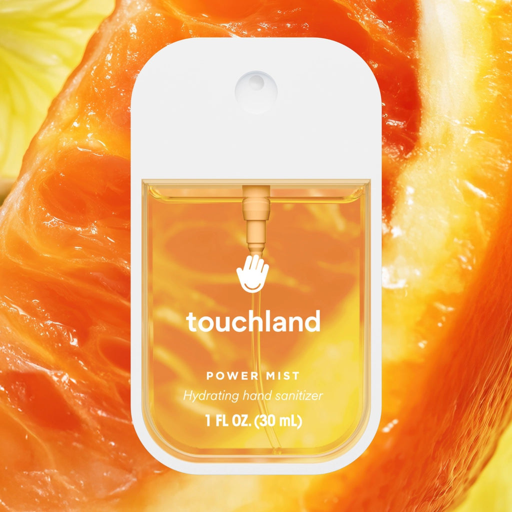 A small Touchland Power Mist hand sanitizer bottle with a white cap and clear front panel showing amber liquid, centered over a close-up orange slice background.