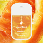 A small Touchland Power Mist hand sanitizer bottle with a white cap and clear front panel showing amber liquid, centered over a close-up orange slice background.