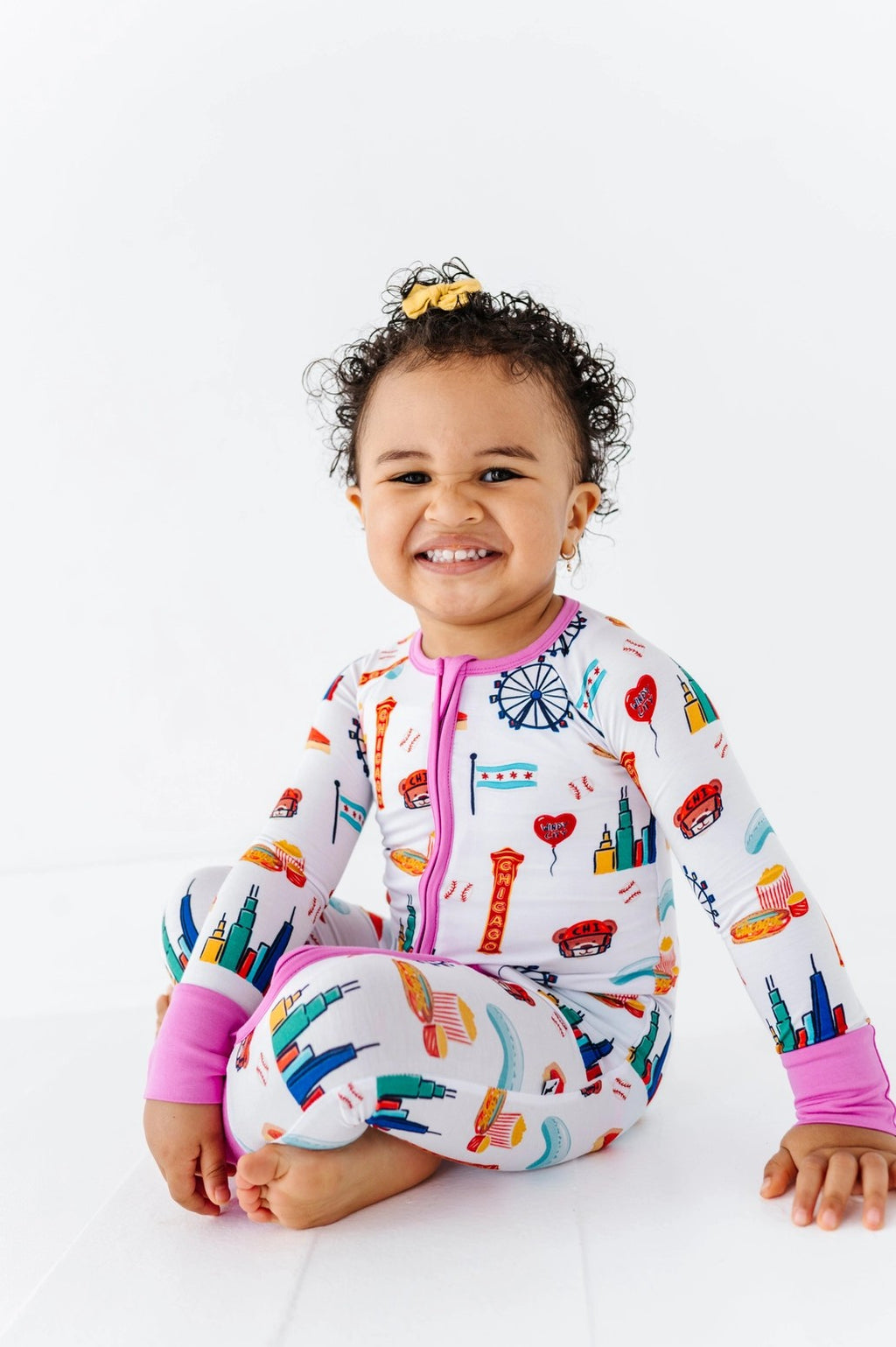 Toddler smiling and sitting while wearing colorful patterned Convertible Footies - Bamboo - Pink Chicago featuring Chicago-themed illustrations and pink trim, cozy and breathable.