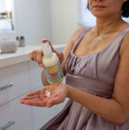 Woman in a soft lavender dress dispensing California Baby - (No Fragrance) Super Sensitive™ Everyday Lotion onto her hand in a bright, cozy bathroom setting, showcasing a soothing skincare moment.