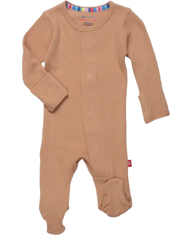 A baby's long-sleeve footed sleeper in a warm tan color, featuring a waffle knit texture, snap closures down the front, and a small red tag on the side.
