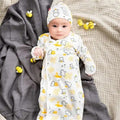A baby lying on a gray blanket and textured beige surface, wearing a long-sleeve white sleep sack and matching bonnet with a yellow and gray bear and honeycomb print.