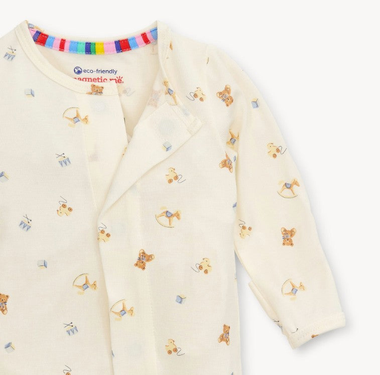 A close-up of a baby long-sleeve romper in cream-colored cotton fabric, featuring a playful print of teddy bears, bows, and arrows in brown, blue, and yellow. The brand 'Commette' is visible on the inner neck label along with 'eco-friendly' and 'cotton' text.