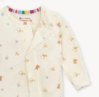A close-up of a baby long-sleeve romper in cream-colored cotton fabric, featuring a playful print of teddy bears, bows, and arrows in brown, blue, and yellow. The brand 'Commette' is visible on the inner neck label along with 'eco-friendly' and 'cotton' text.