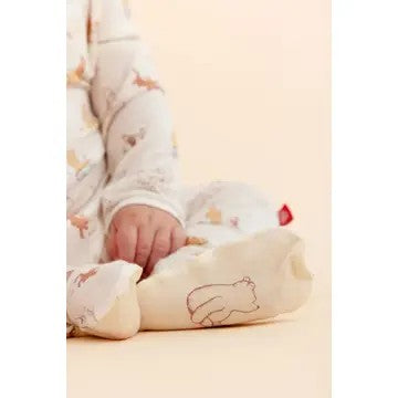 A close-up of a baby wearing a cream-colored footed sleeper with Winnie the Pooh print, resting on a matching cream pillow with a bear outline.