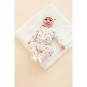 A baby wearing a cream-colored footed sleeper with Winnie the Pooh print and a matching beanie hat, lying on a white blanket against a pale peach background.