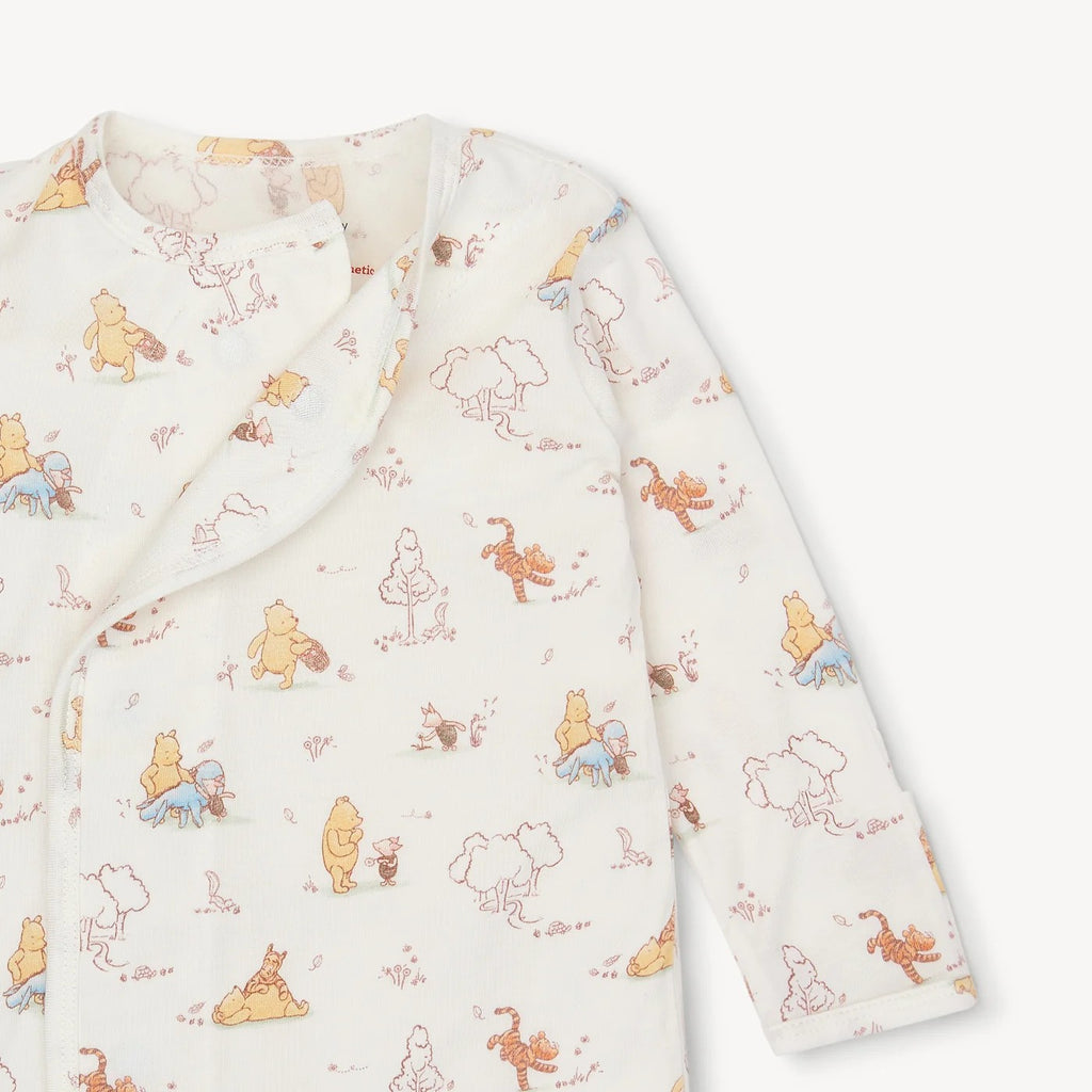 A long-sleeve baby sleep sack or pajama top in a warm off-white fabric, featuring a repeating print of Winnie the Pooh and his friends in soft pastel colors.