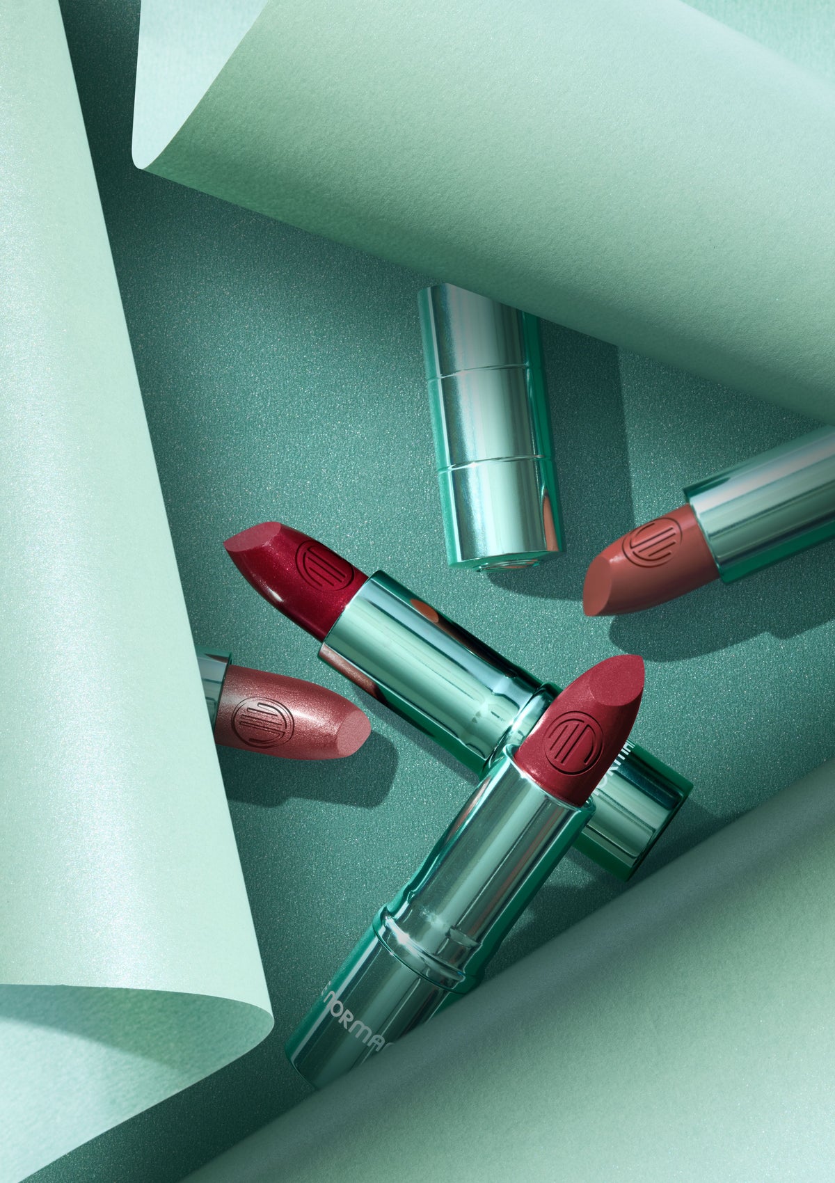 Four lipstick tubes arranged diagonally on a mint green surface with matching paper-like corner props. The lipsticks have metallic teal-green cases and show lipstick bullets in red, pink, and nude tones.