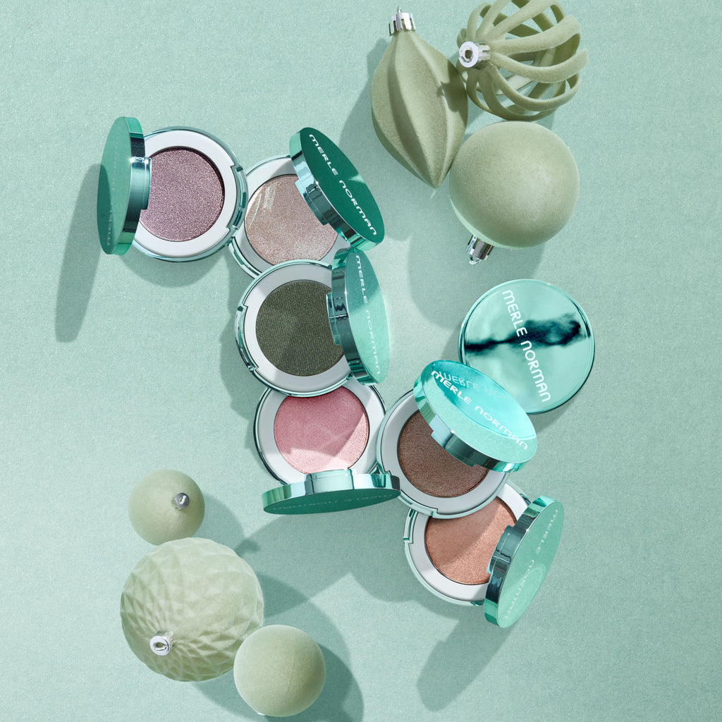 An overhead flat-lay of several open round makeup compacts with mint green lids and shimmering pressed powders in pink, taupe, olive, and peach tones, arranged with matching mint-green decorative ornaments on a pale mint background. The compacts show the brand name Merle Norman.