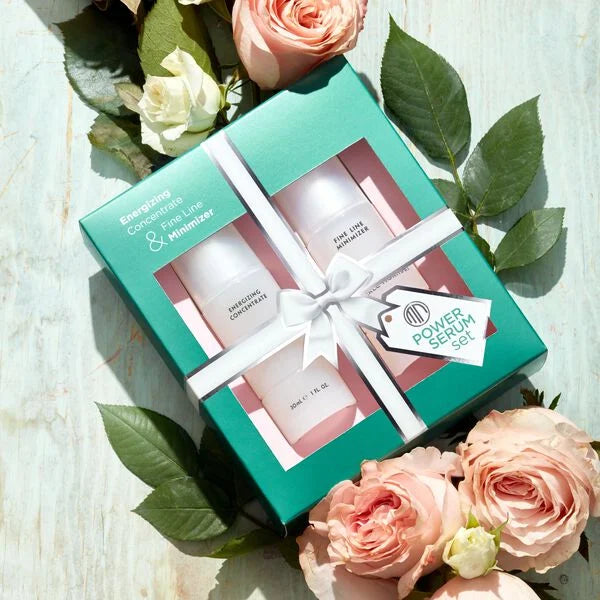 Merle Norman Cosmetics - Power Serum Set (Limited-Edition) displayed in teal packaging with white ribbon, surrounded by soft pink and white roses on a light wooden surface.