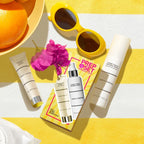 Flat lay of the Prep & Set Makeup Duo (Limited-Edition) with sunscreen, setting spray, yellow sunglasses, and vibrant bougainvillea on a yellow and white striped surface, styled for mom life essentials.