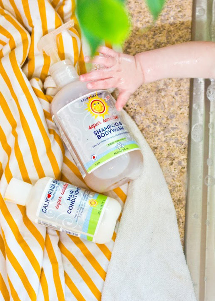Baby’s hand reaching for a bottle of California Baby - (No Fragrance) Super Sensitive™ Hair Conditioner next to shampoo and body wash on a soft striped towel near a sink.