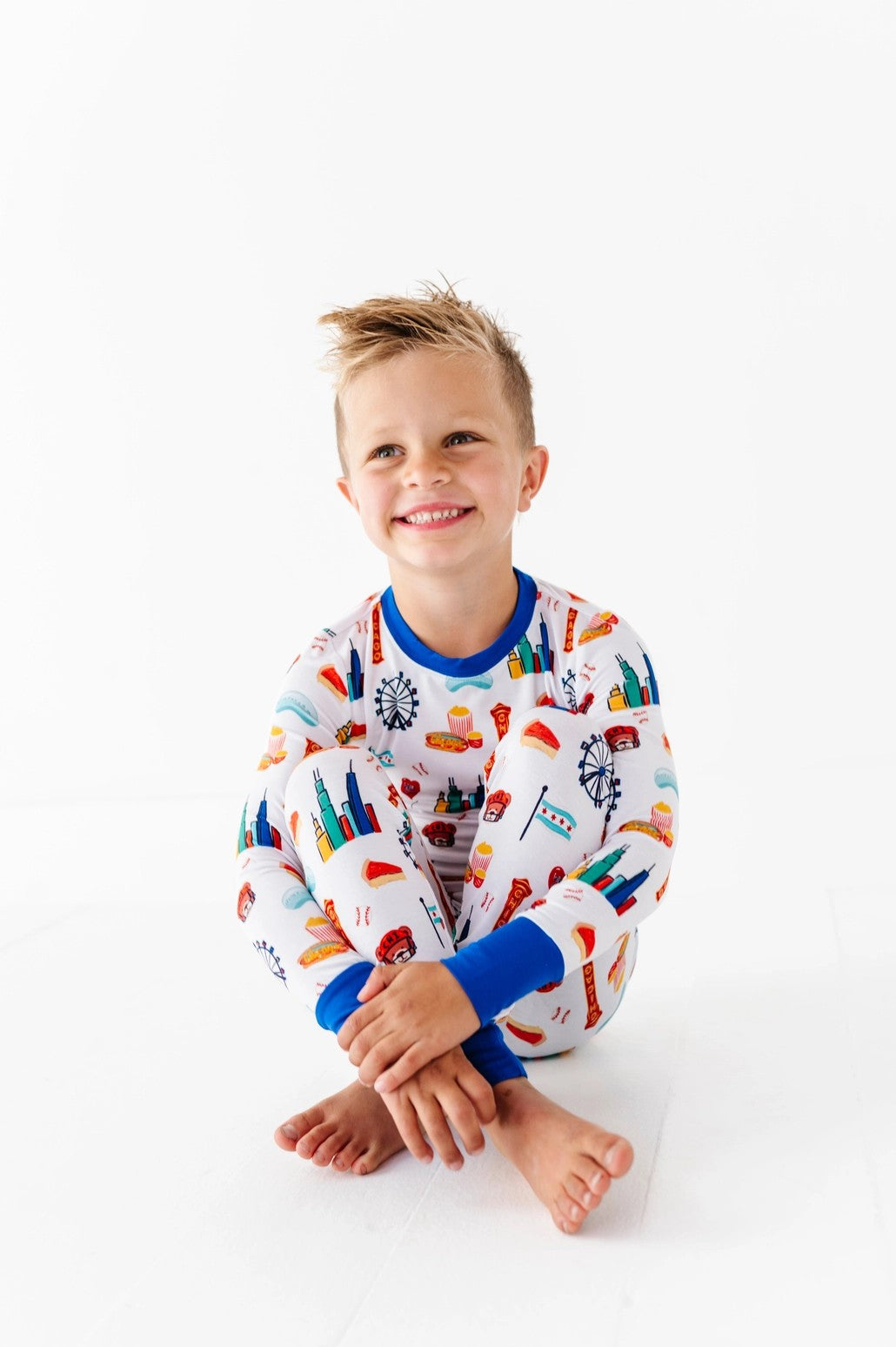 Smiling young boy sitting cross-legged wearing Two-Piece Pajamas - Bamboo - Blue Chicago featuring colorful Chicago-themed prints with blue cuffs, styled for cozy comfort and practical mom essentials.