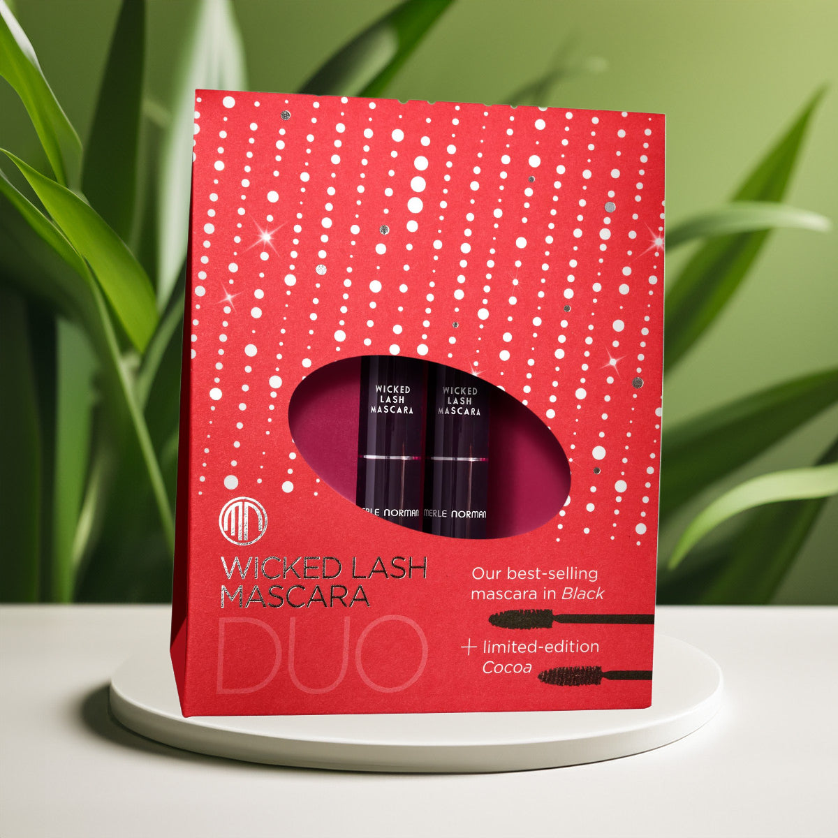 Merle Norman Cosmetics - Wicked Lash Mascara Duo (Limited-Edition) in festive red packaging featuring best-selling black and limited-edition cocoa mascaras, styled with natural greenery.