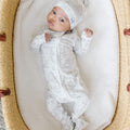 Baby swaddled in a white blanket with patterns, wearing a matching hat, in a woven basket.