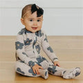 A baby girl sitting on a light wood floor wearing a cream-colored footed one-piece pajama with a blue-gray floral pattern and a matching headband with a black bow.