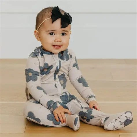 A baby girl sitting on a light wood floor wearing a cream-colored footed one-piece pajama with a blue-gray floral pattern and a matching headband with a black bow.