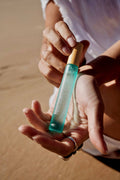 Hand holding a small turquoise and gold bottle of True Ocean - True Ocean Body Spray - Pocket Size with a sandy background, capturing a natural and stylish moment.