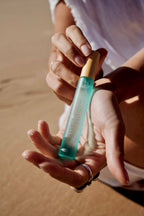 Hand holding a small turquoise and gold bottle of True Ocean - True Ocean Body Spray - Pocket Size with a sandy background, capturing a natural and stylish moment.