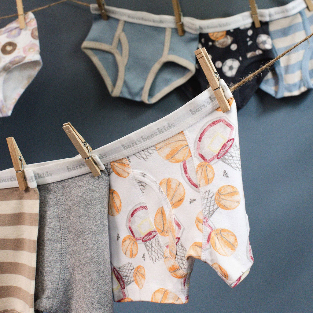 A close-up of children's boxer briefs hanging on a clothesline with wooden clothespins against a dark blue-gray wall. The front pair is white with a basketball and net pattern, with gray and striped pairs visible behind it.