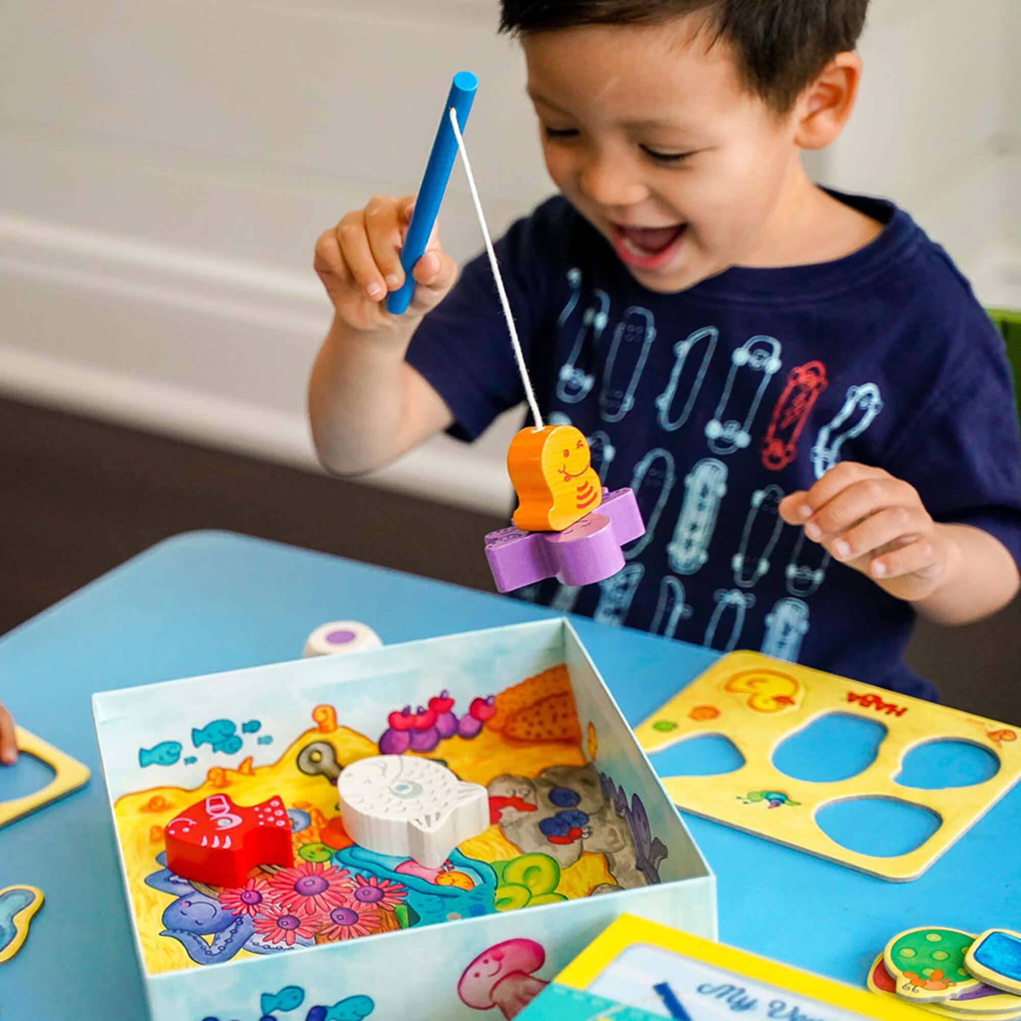 A young child playing with the "My Very First Games - Here, Fishy, Fishy! Magnetic Game," using a magnetic fishing rod to catch colorful wooden fish on a bright play table, promoting imaginative learning and sensory play.