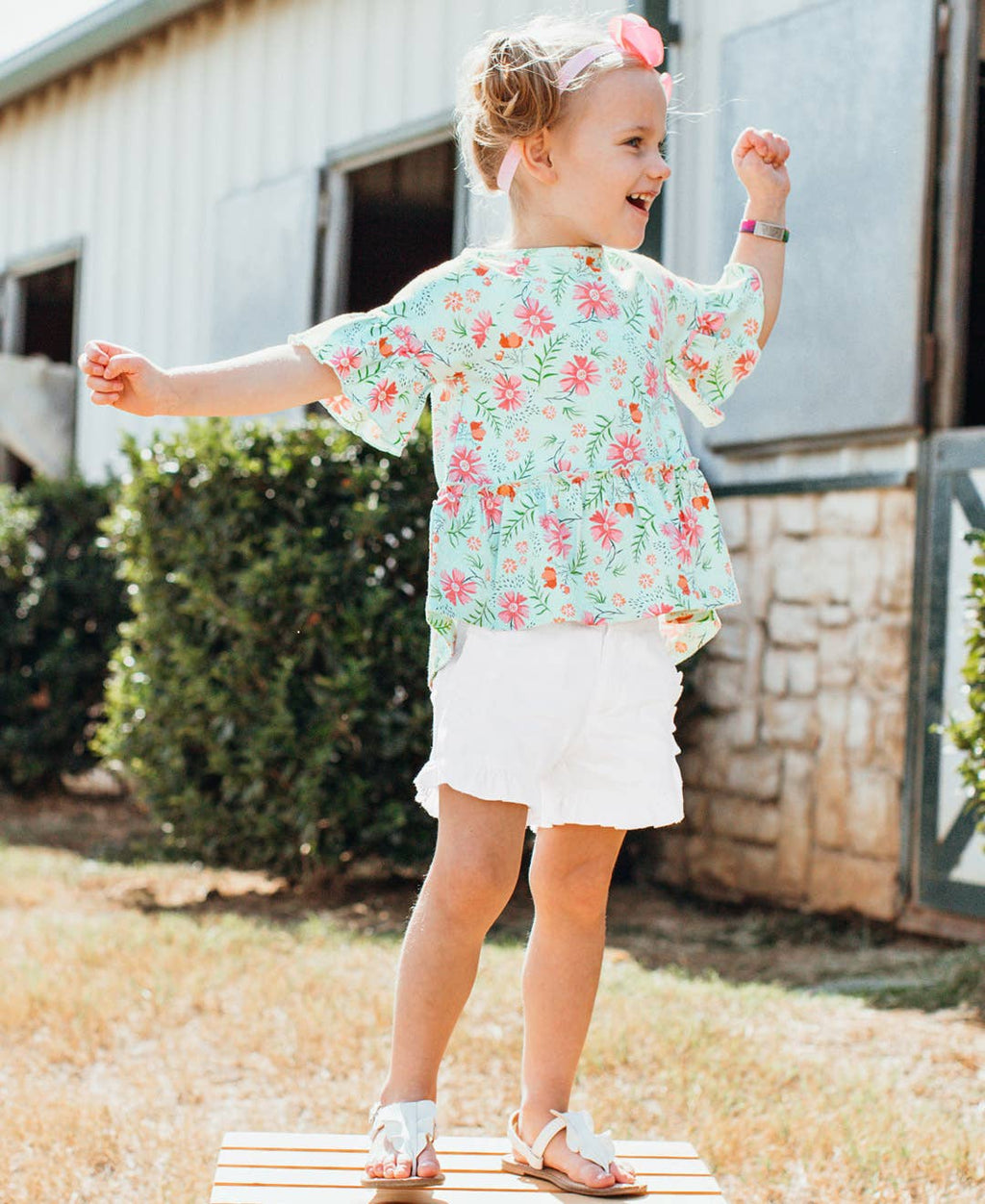 Toddler girl wearing RuffleButts + RuggedButts - Toddler Girls White Ruffle Trim Woven Shorts: White, paired with a floral top, playing outdoors in a sunny garden setting.
