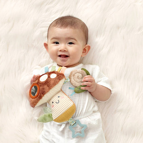 Happy baby lying on a soft white blanket holding the Itzy Ritzy - Bitzy Traveler™: Mushroom plush toy designed for sensory play and comfort with soothing neutral colors.