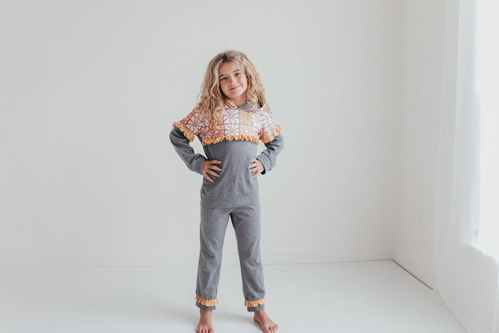 A young girl stands barefoot in a bright, minimalist white room, wearing a gray long-sleeve jumpsuit with a colorful patterned ruffled collar and orange ruffled trim at the cuffs.