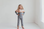 A young girl stands barefoot in a bright, minimalist white room, wearing a gray long-sleeve jumpsuit with a colorful patterned ruffled collar and orange ruffled trim at the cuffs.