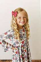 Young girl wearing a floral dress standing on a wooden floor against a white wall.