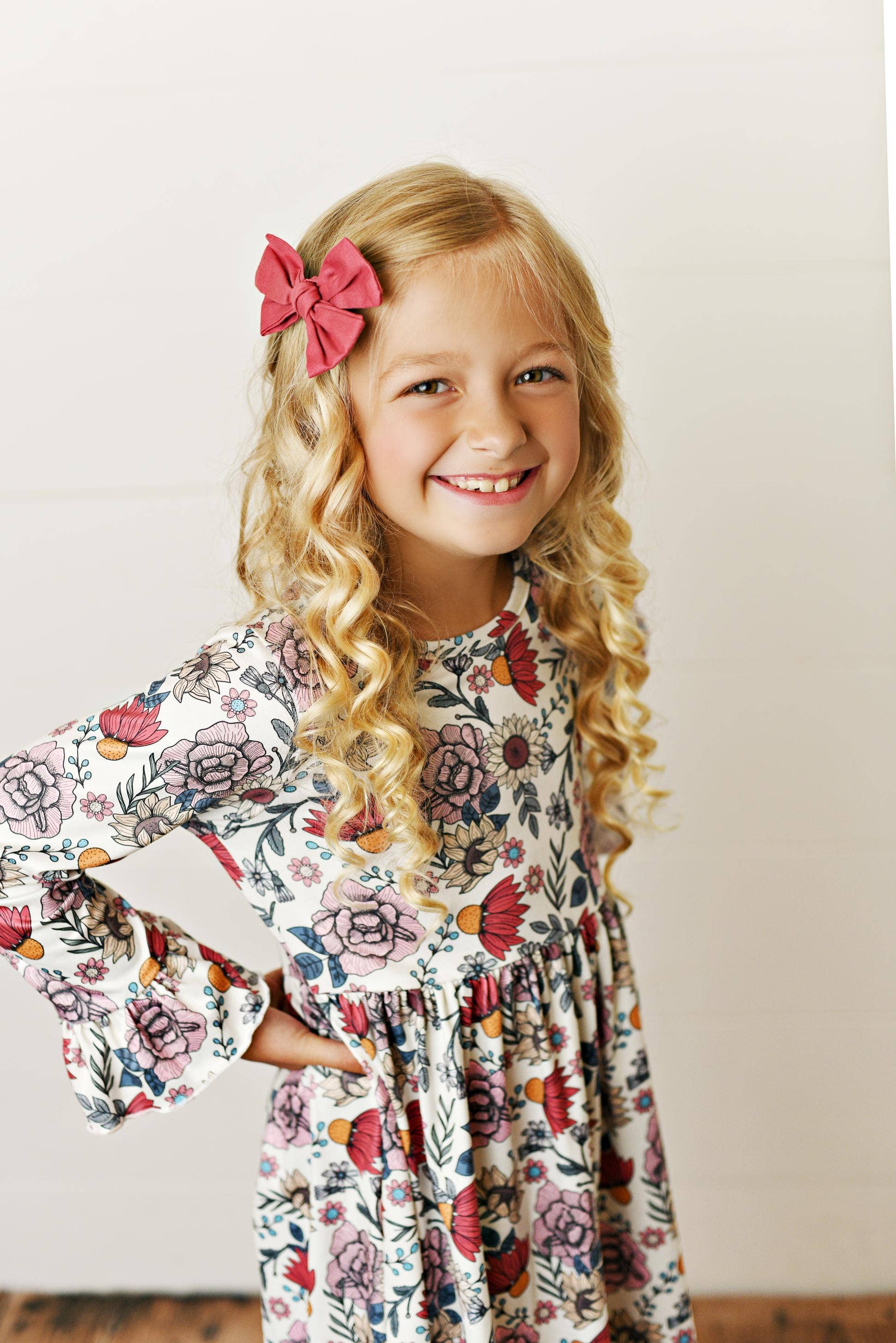 Young girl wearing a floral dress standing on a wooden floor against a white wall.
