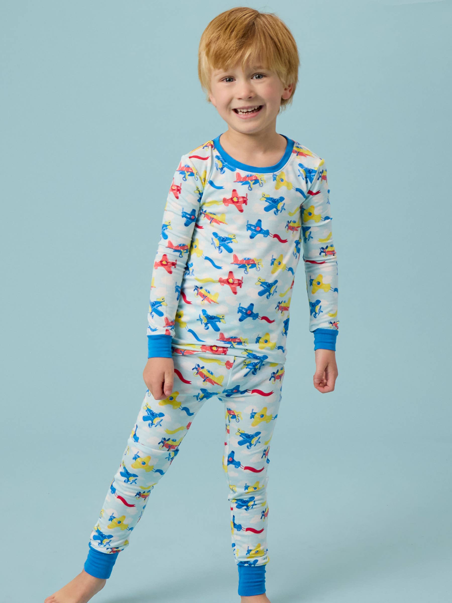 Smiling young boy wearing the RuffleButts + RuggedButts - SoftSnooze™ Kids Bamboo Viscose Plane Parade Long Sleeve Pajama Set featuring colorful airplanes on a light background, styled for comfort and playful bedtime.