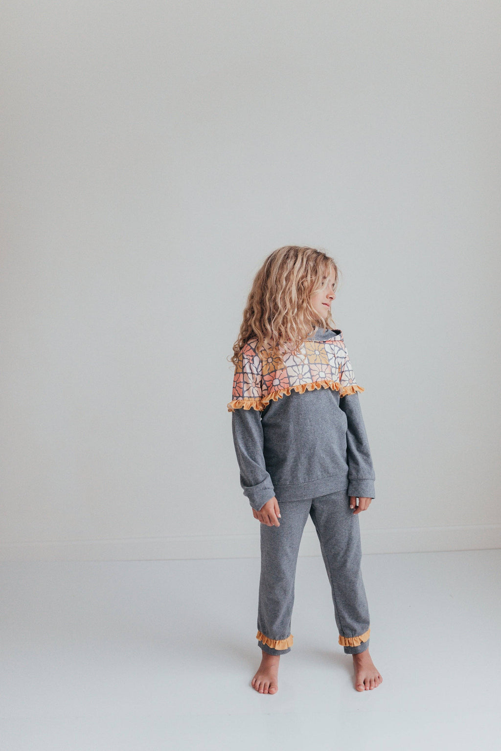 A young girl with long blonde hair stands barefoot in a studio, wearing a gray two-piece pajama set with a colorful geometric patterned yoke and orange pom-pom trim.