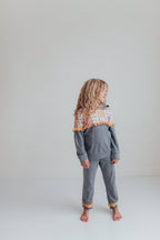 A young girl with long blonde hair stands barefoot in a studio, wearing a gray two-piece pajama set with a colorful geometric patterned yoke and orange pom-pom trim.