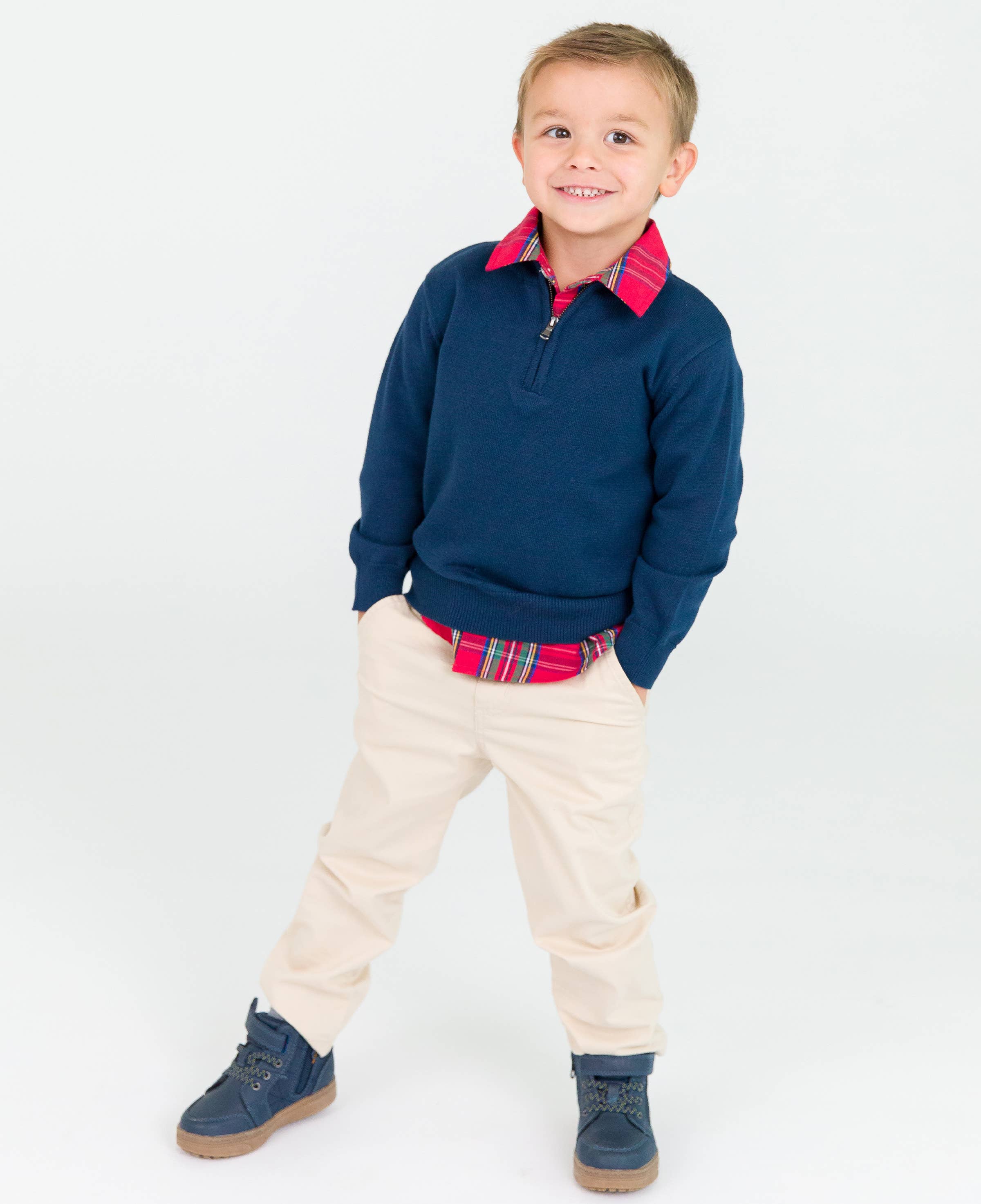 Young boy smiling confidently wearing RuffleButts + RuggedButts - Boys Khaki Stretch Chino Pants: Off-White with navy sweater, red plaid shirt, and navy boots, styled for comfortable, modern kids fashion.