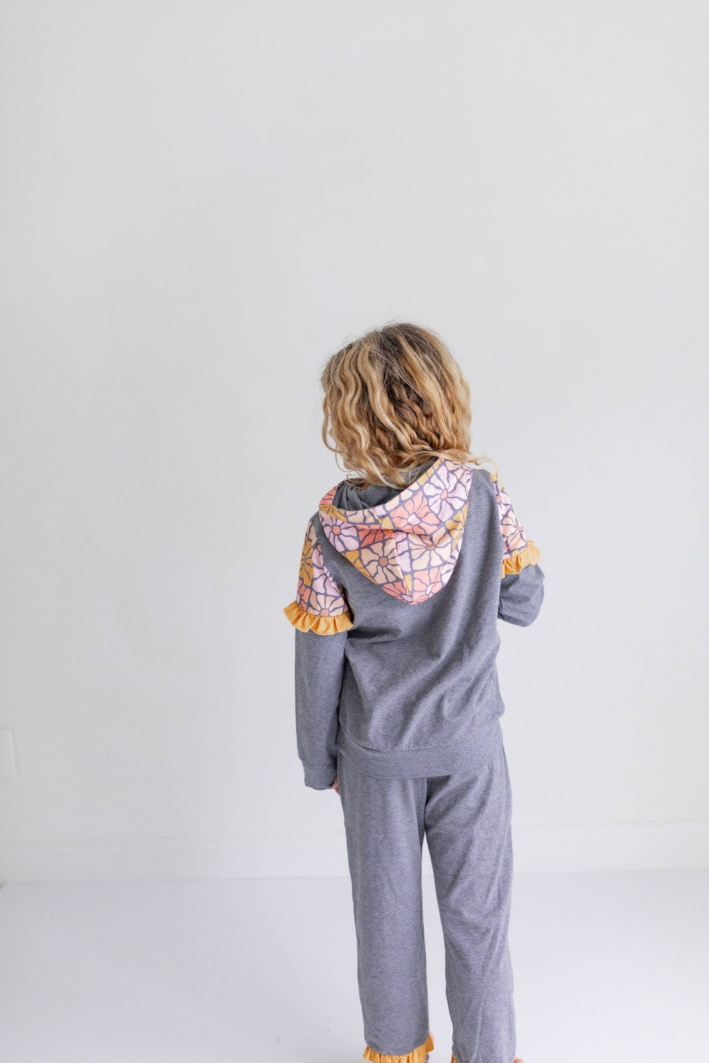 A child standing with their back to the camera, wearing a gray hooded sweatshirt and matching sweatpants. The hoodie features a colorful geometric patterned hood lining and yellow ruffle trim along the sleeves and cuffs.