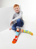 A smiling child wearing Pals Socks - Taco & Hot Sauce Mismatched Non-Slip Socks for Kids, featuring bright colorful taco and hot sauce designs, styled with casual jeans and a gray sweater.