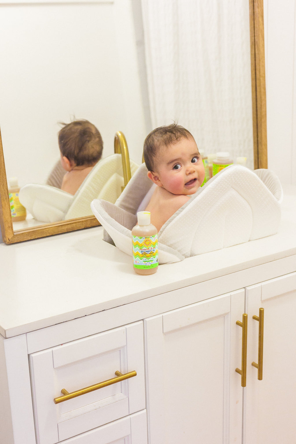 Baby sitting in a white cushioned bath seat on a white vanity countertop next to a bottle of California Baby - Chamomile & Mint™ Shampoo & Bodywash, with a mirror and gold hardware in the background.