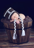 Newborn baby peacefully sleeping in a wooden bucket, wearing The Daisy Baby LLC - Brown/White Football Crocheted Handmade Heirloom Gift Hat, styled for cozy baby boutique photography.