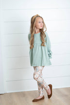 Young girl wearing a light green dress and floral leggings against a white wooden panel background