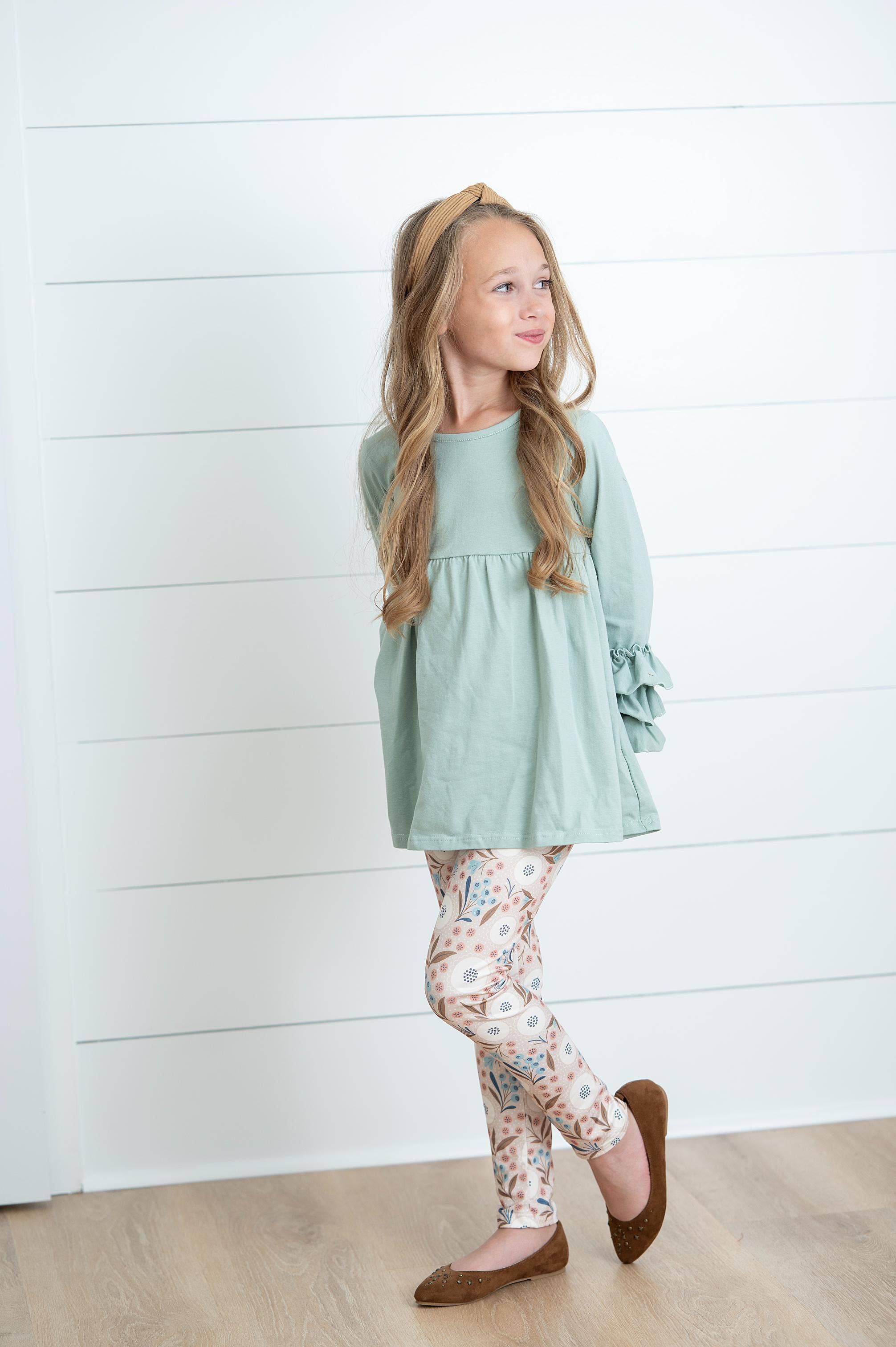 Young girl wearing a light green dress and floral leggings against a white wooden panel background