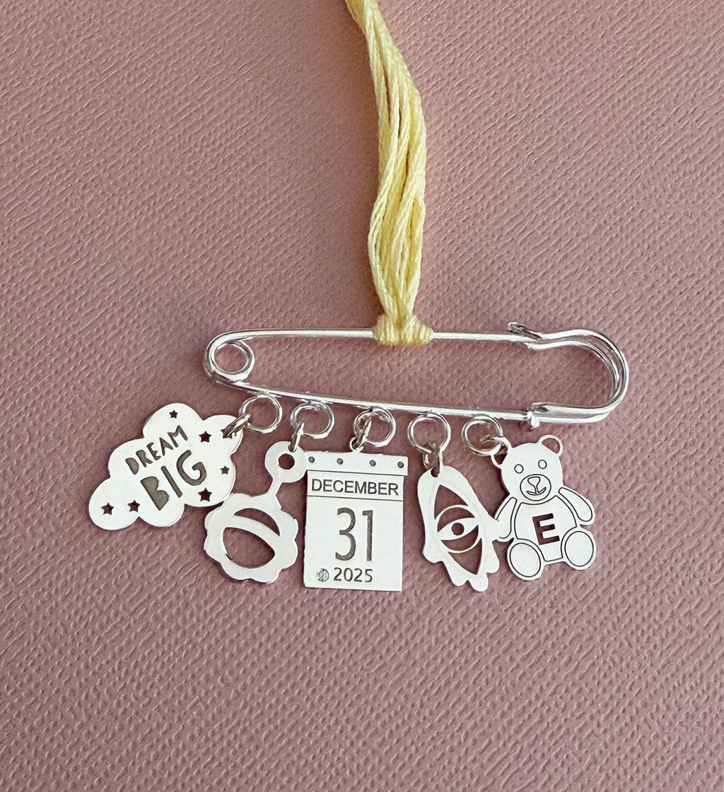 Silver safety pin charm featuring personalized hanging charms including a "Dream Big" cloud, baby face, calendar date December 31 2025, rocket, and teddy bear with initial E, designed by Miriam Merenfeld Jewelry - Personalized Safety Pin, styled for modern moms and children’s boutique shoppers.