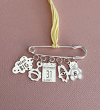 Silver safety pin charm featuring personalized hanging charms including a "Dream Big" cloud, baby face, calendar date December 31 2025, rocket, and teddy bear with initial E, designed by Miriam Merenfeld Jewelry - Personalized Safety Pin, styled for modern moms and children’s boutique shoppers.
