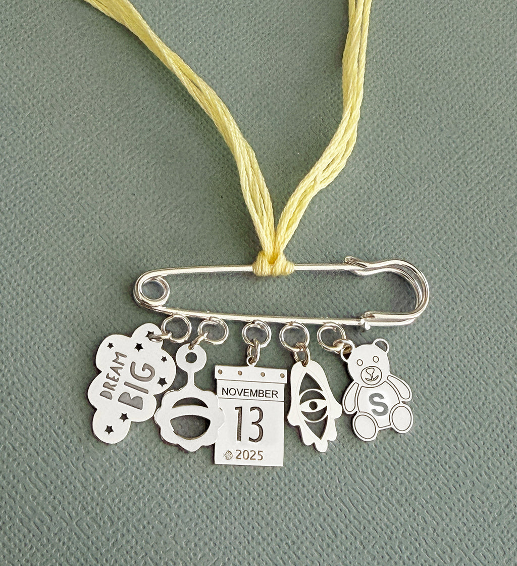 Silver safety pin pendant with hanging charms including "Dream Big" cloud, pacifier, calendar date, eye symbol, and teddy bear on a yellow cord, Miriam Merenfeld Jewelry - Personalized Safety Pin.