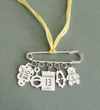 Silver safety pin pendant with hanging charms including "Dream Big" cloud, pacifier, calendar date, eye symbol, and teddy bear on a yellow cord, Miriam Merenfeld Jewelry - Personalized Safety Pin.