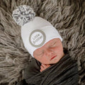 Newborn baby peacefully sleeping while wearing the ilybean - White Little Brother Mixed Gray Pom Newborn Hospital Hat, wrapped in a cozy dark gray blanket on a soft textured surface.