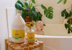 California Baby - Calendula™ Shampoo + Conditioner Bundle displayed on a wooden stool next to a bathtub with a happy baby and green plants in the background, showcasing cozy baby essentials and mom life essentials.