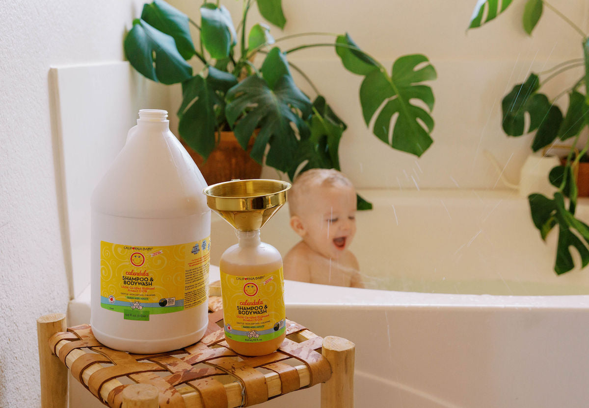 California Baby - Calendula™ Shampoo + Conditioner Bundle displayed on a wooden stool next to a bathtub with a happy baby and green plants in the background, showcasing cozy baby essentials and mom life essentials.