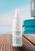 Bottle of SUNPLUS - After Sun Skin Soothing Spray with aloe vera, argan, and vitamin E placed on textured wooden surface by a pool under soft daylight, promoting soothing hydration.